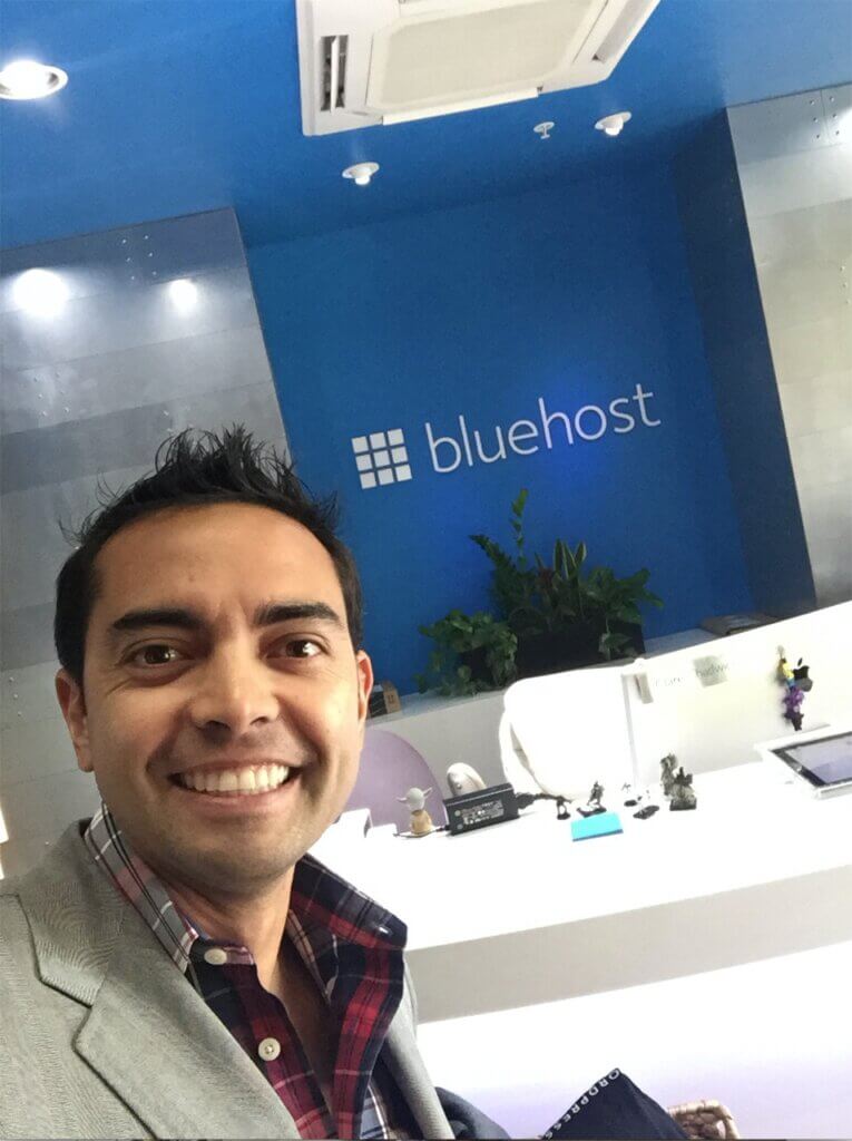 Pat Flynn stands in the Bluehost lobby for a self. A white reception desk and a big blue wall with the word "Bluehost" is behind him.