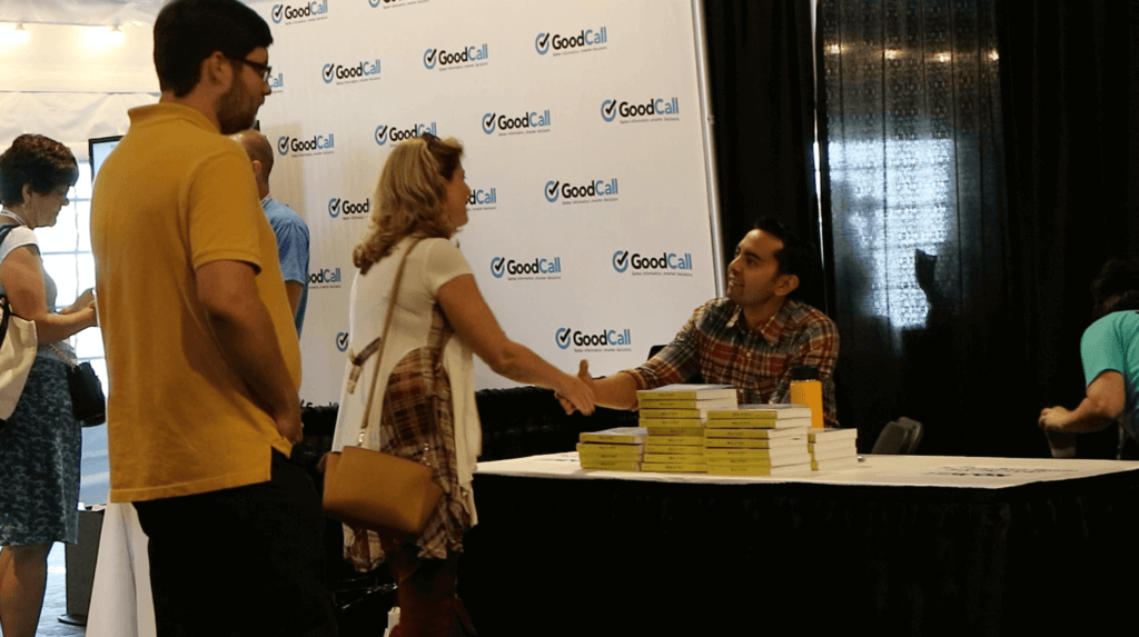 Picture of Pat at a conference table, signing books and shaking a woman's hand
