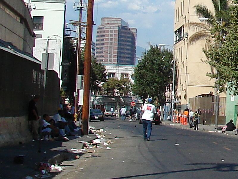 A photo of Skid Row, a street cutting behind tall buildings, with lots of trash on the street and people sitting on the sidewalks.