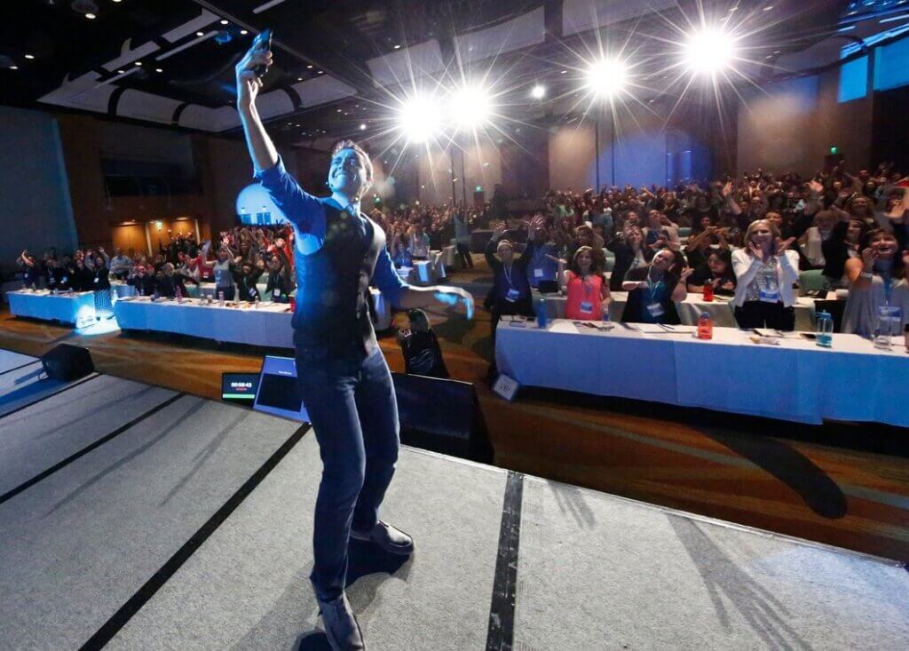 Pat taking a selfie while onstage at the Smart Success Summit. A large audience seated at long tables watches.