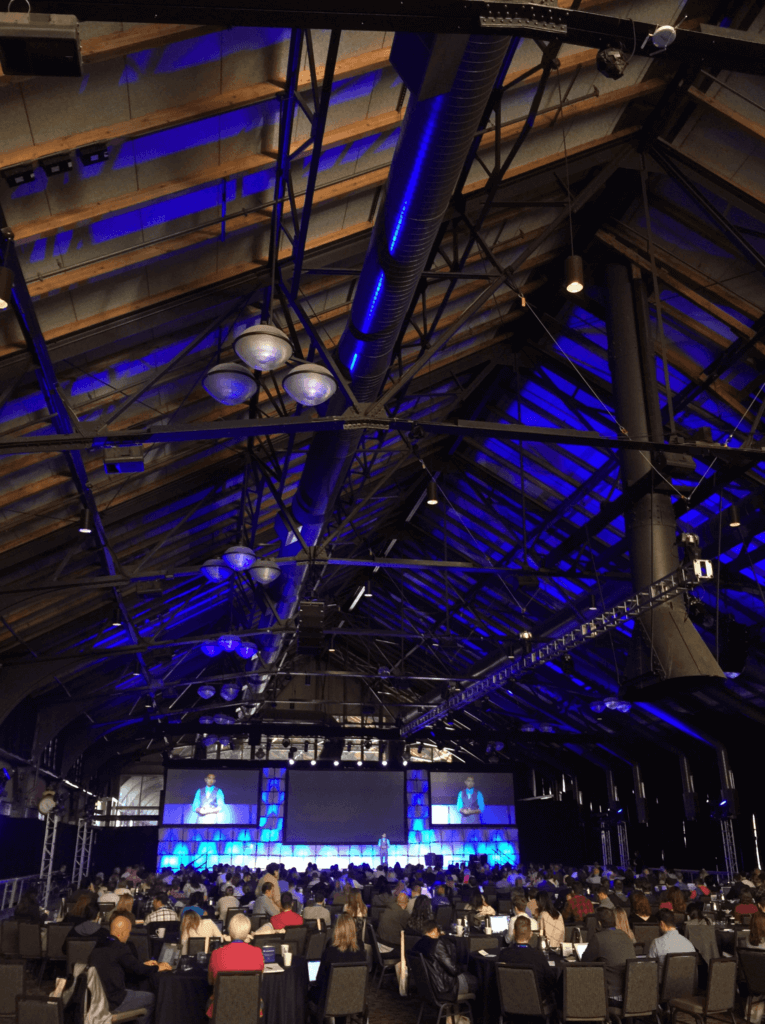 Picture of large conference center with open rafters. Pat is on stage, people are seated at round tables, and the video monitors over the stage show Pat speaking.