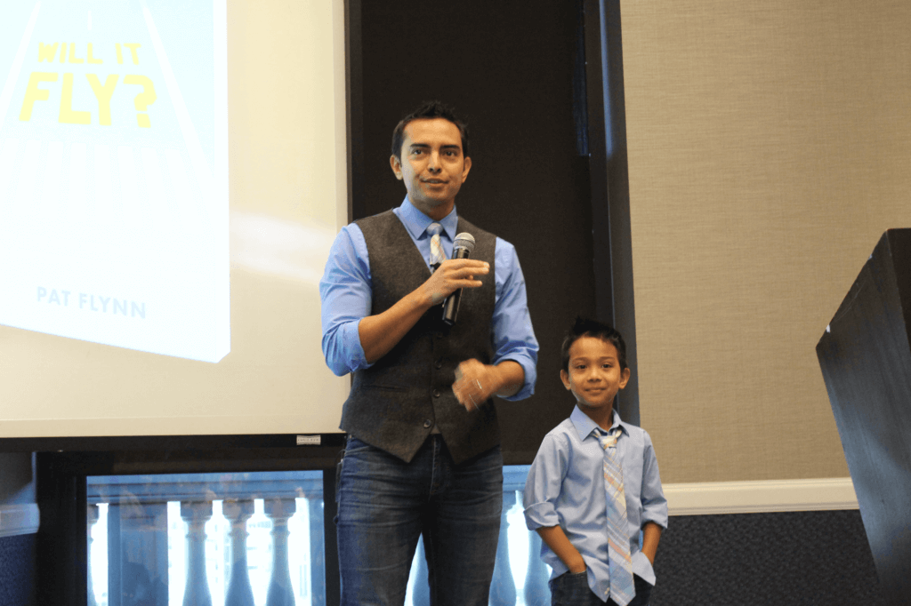 Picture of Pat speaking with a microphone in front of a group. His young son Keoni is with him, dressed in a shirt and tie like his dad.