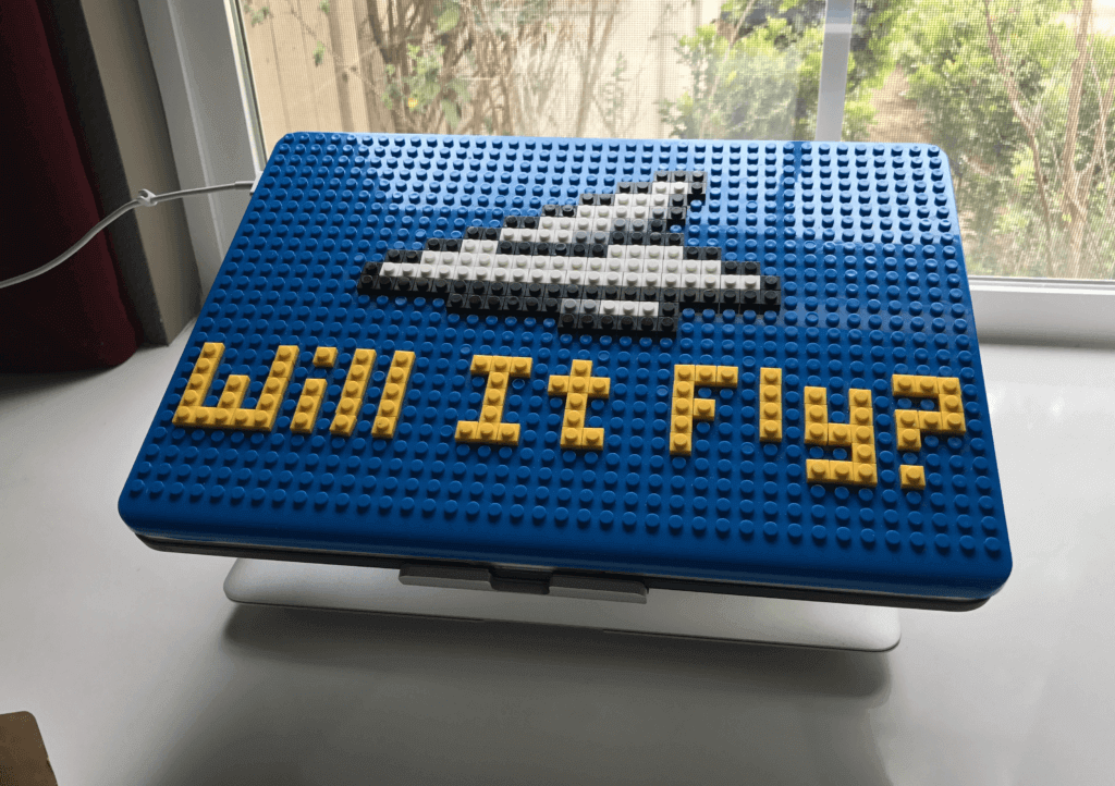 Blue laptop case is also a LEGO base. Pat has flat LEGO spelling out "Will It Fly?" and making a paper airplane.