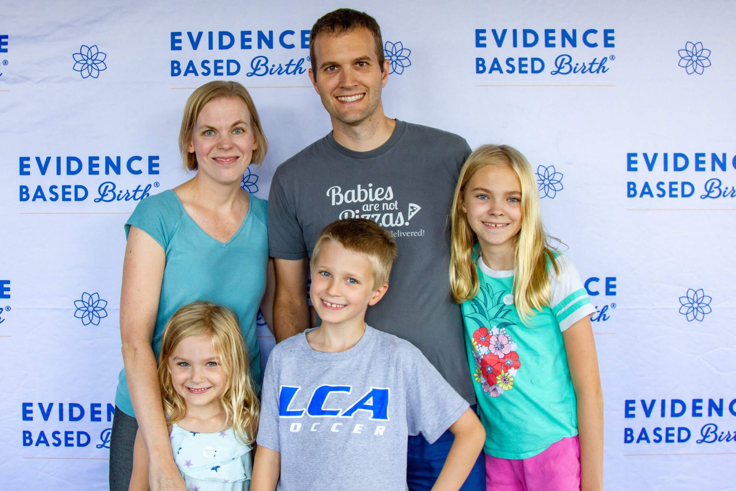 Rebecca Dekker, her husband, and three kids (two girls and one boy) posing in front of a step and repeat that says "Evidence Based Birth"