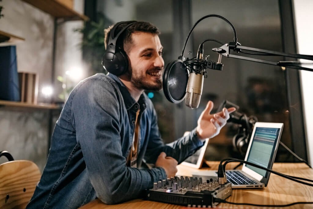 man wearing headphones smiling as he talks into a microphone with pop filter at a desk with a laptop and mixer, recording a guest podcasting episode 