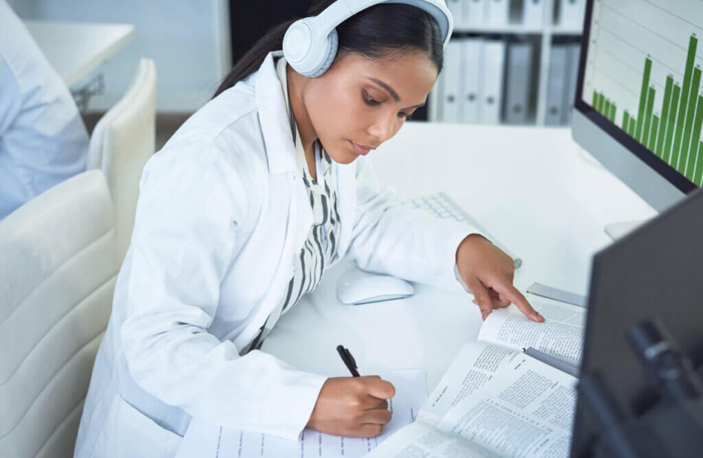 woman sitting at desk by computer in white lab coat listening to headphones while reading from a book and writing notes
