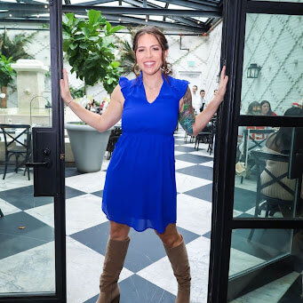 Katy Widrick poses in a blue dress and cowboy boots in the doorway of a brightly-lit glass room with plants.