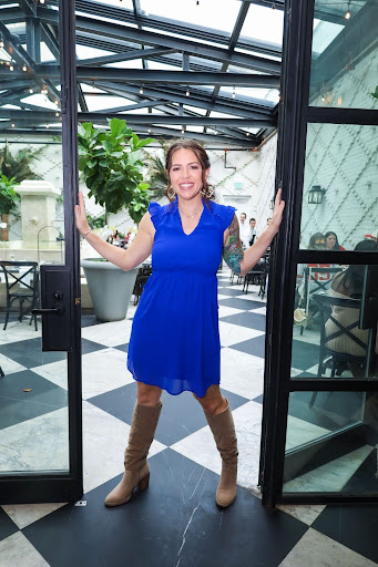 Katy Widrick poses in a blue dress and cowboy boots in the doorway of a brightly-lit glass room with plants.