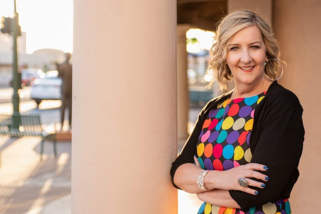 Pamela Slim standing next to a pillar, wearing a colorful blouse, and smiling at the camera.